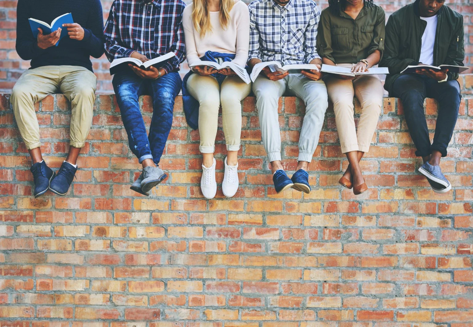 Gruppo di giovani seduti su un muro di mattoni che leggono libri insieme.